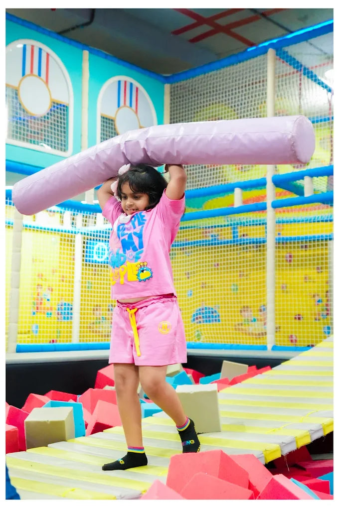 A young girl in pink shorts and t-shirt lifts a large purple foam cylinder overhead in a colorful indoor play area.