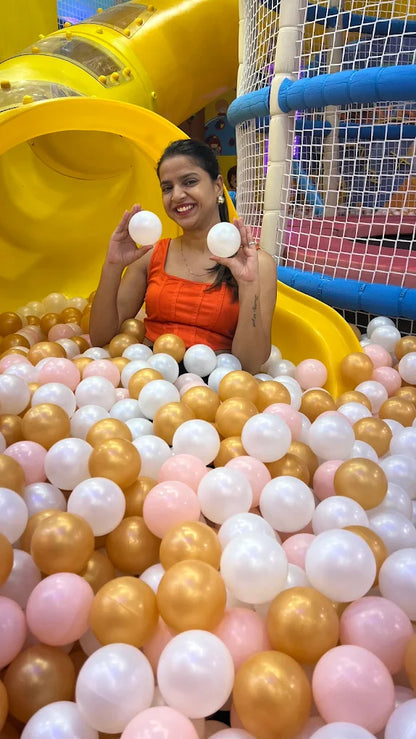A young woman smiles while holding two white balls in a ball pit with a yellow slide.