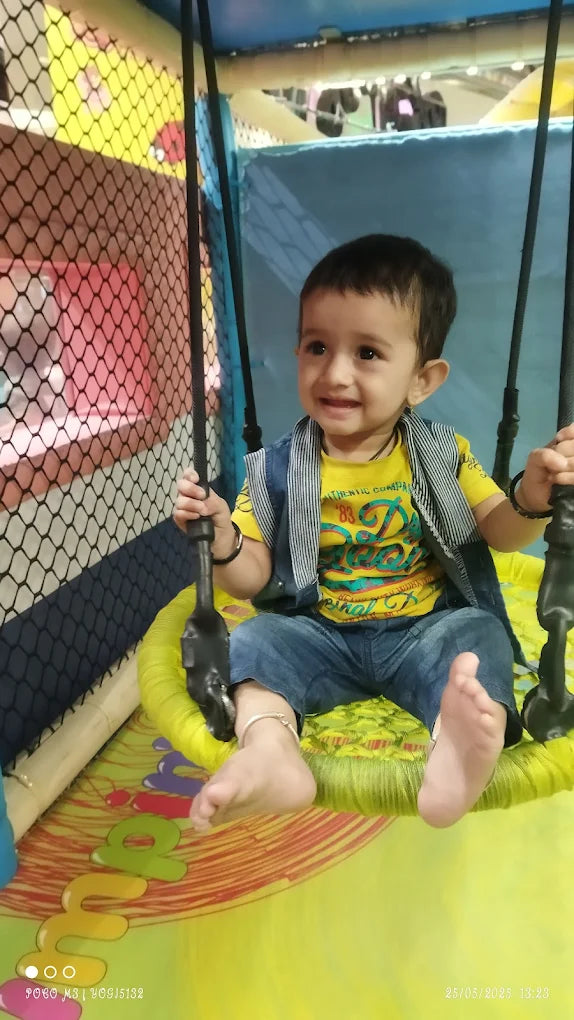 A smiling toddler sits on a yellow swing in a play area. The child wears a yellow shirt and denim vest.