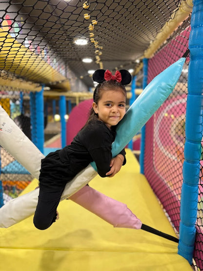 A young girl in Minnie Mouse ears smiles while lying on a padded structure in a colorful indoor playground.