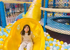 A young child smiles from a yellow slide in a ball pit at an indoor playground.