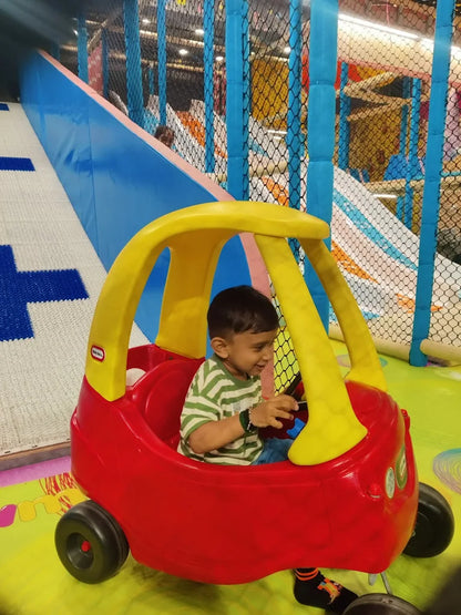 A young child sits in a red toy car in a colorful indoor playground. The child is smiling and holding the steering wheel.
