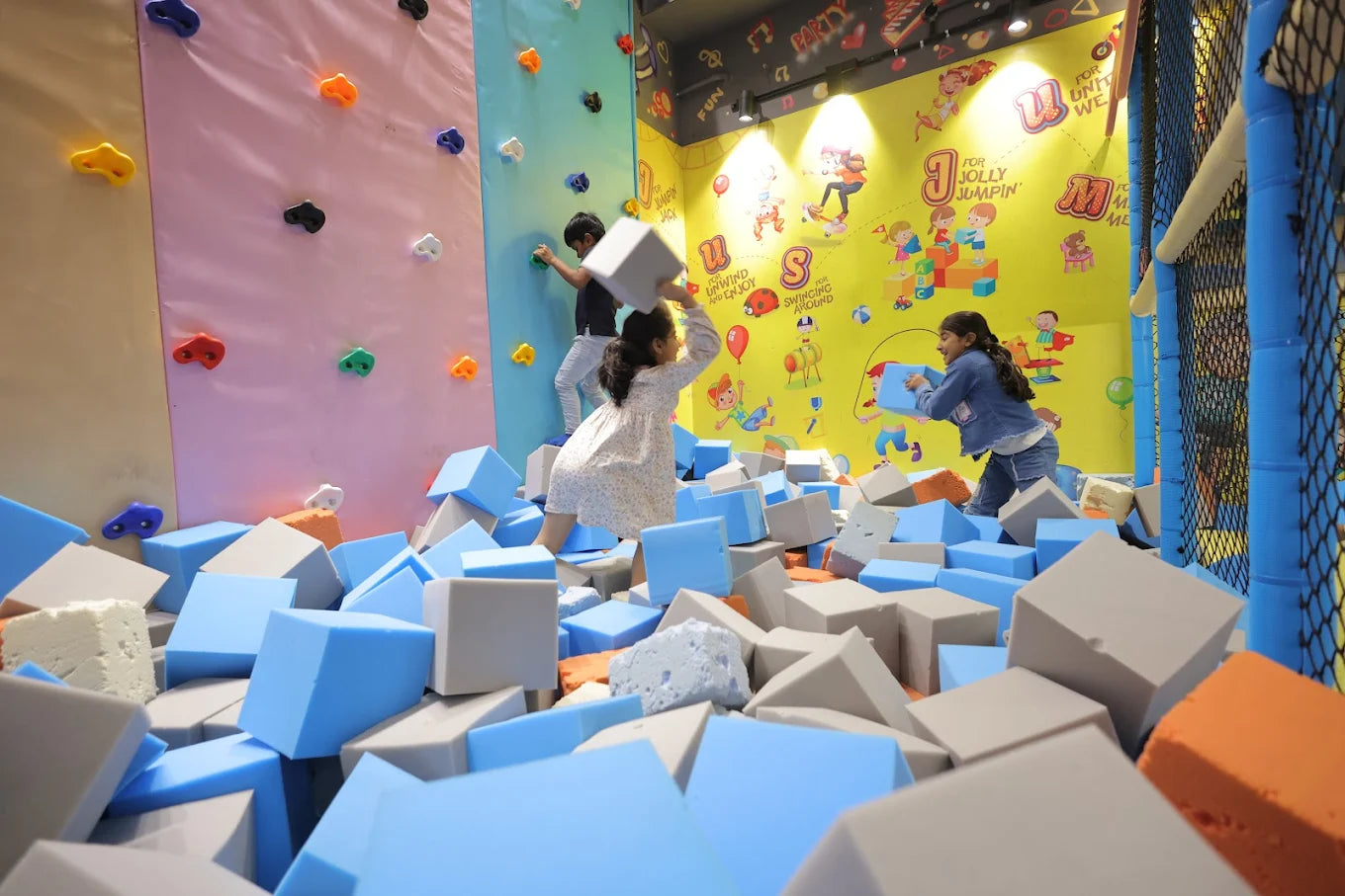 Children play in a foam pit with a climbing wall and colorful murals in the background.