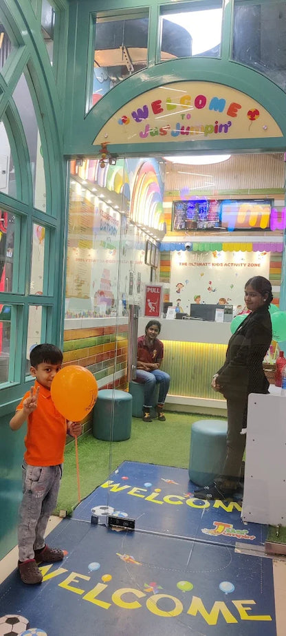 A young boy holds an orange balloon and makes a peace sign in front of a "Welcome to Jus Jumpin'" sign. Two women are visible in the background of the indoor play area.