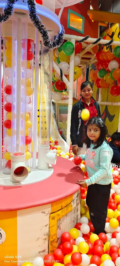 A young girl plays with red balls in a ball pit, with a woman standing behind her in a colorful indoor playground.