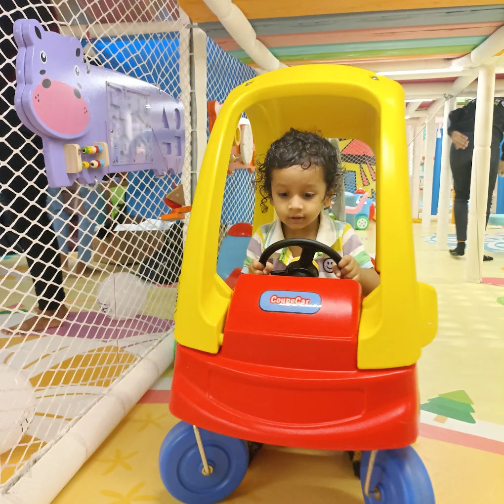 A young child sits in a red and yellow toy car in a colorful indoor play area.
