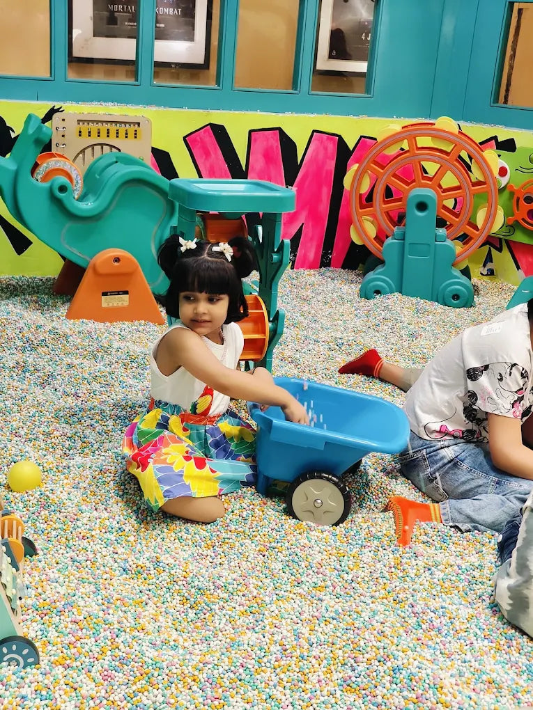 A young child plays with a blue wheelbarrow in a ball pit filled with colorful balls.