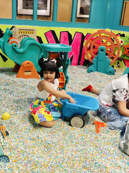 A young child plays with a blue wheelbarrow in a ball pit filled with colorful balls.