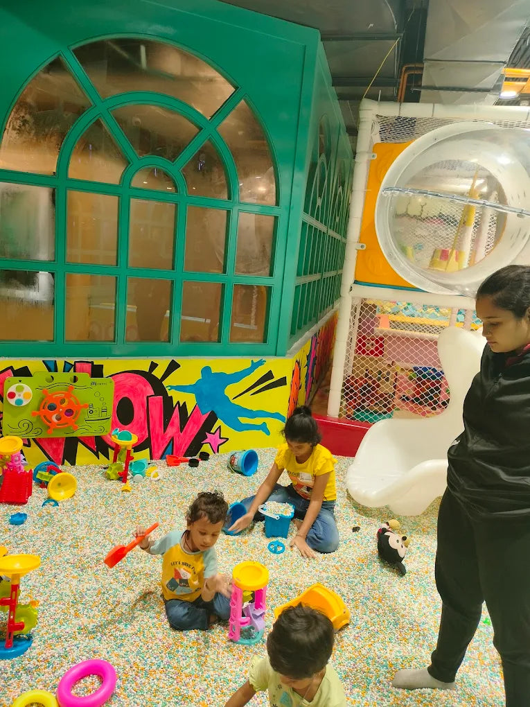 Children play in a ball pit with toys, while a woman supervises.
