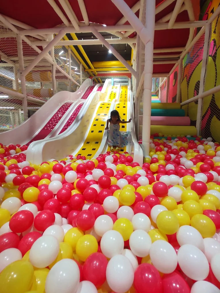A young girl plays in a colorful ball pit next to a slide in an indoor playground.