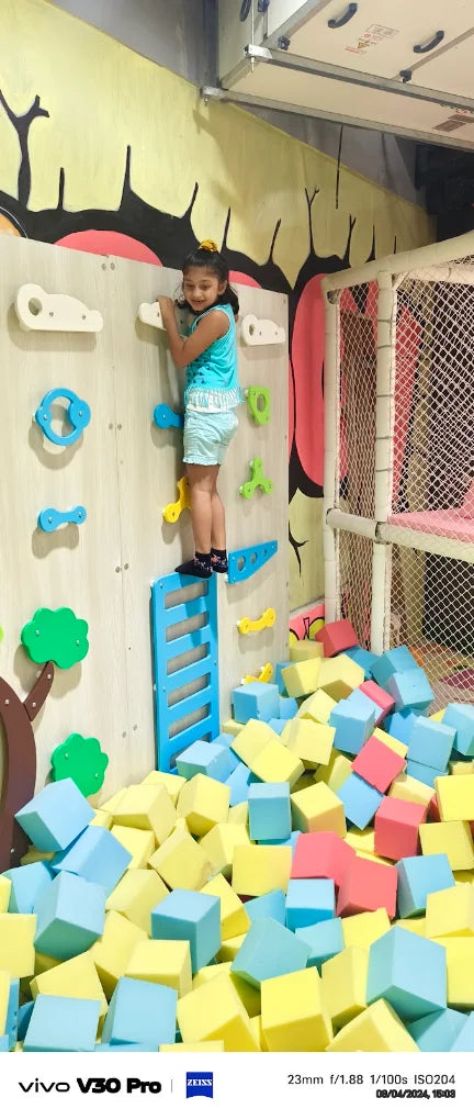 A young girl climbs a rock wall above a ball pit filled with colorful foam cubes.