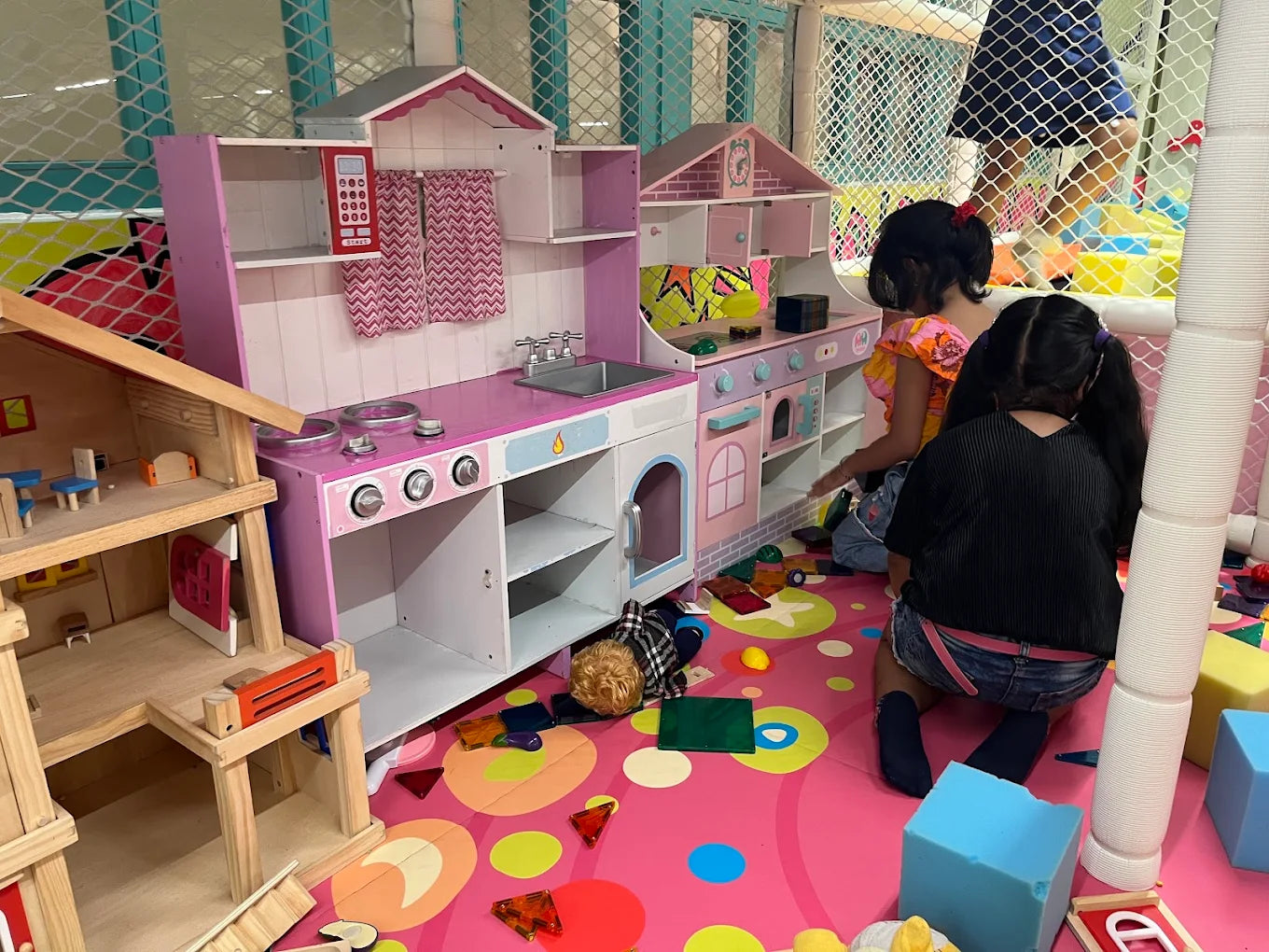 Two young girls play with a toy kitchen and dollhouse in a colorful indoor play area.