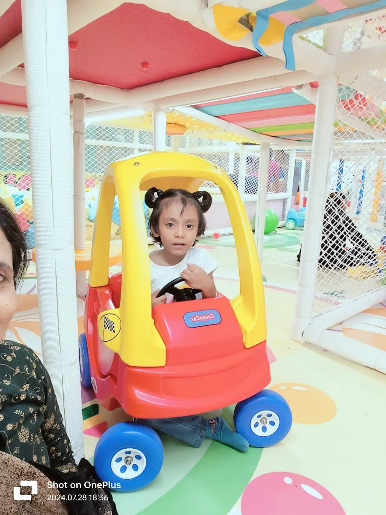 A young child sits in a red and yellow toy car in a colorful indoor playground.