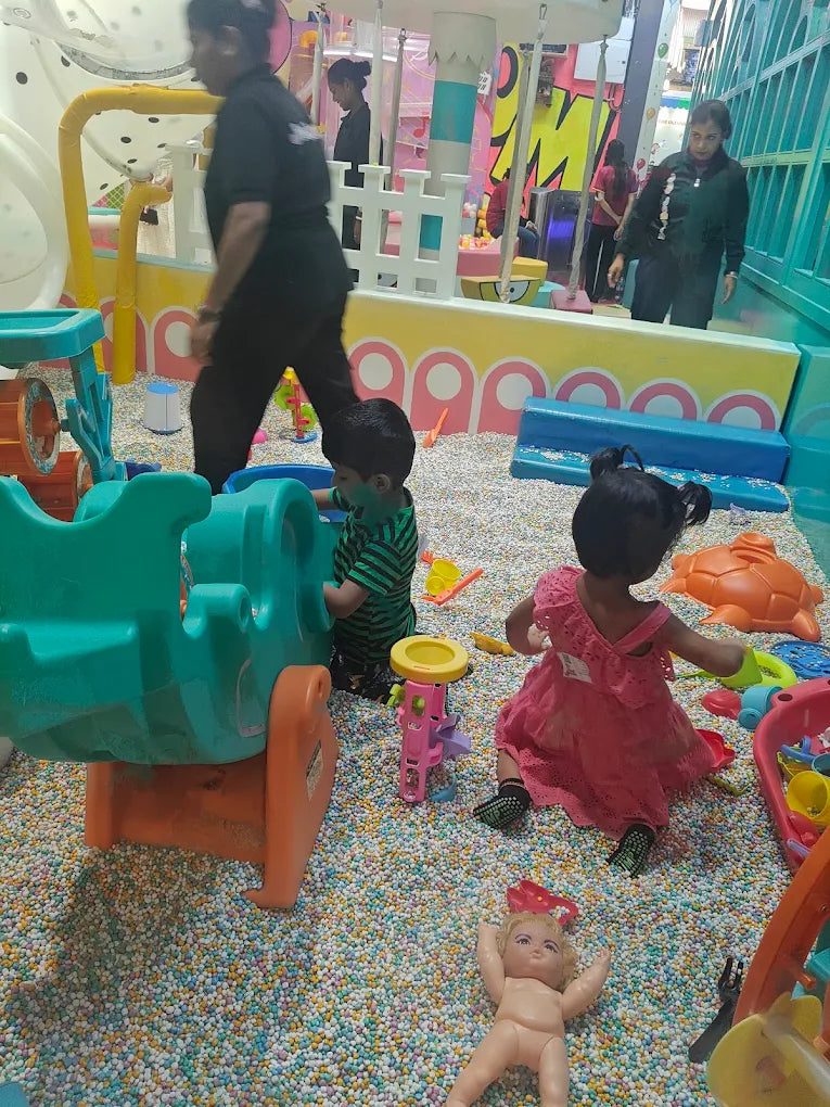 Children play in a colorful ball pit with toys. Adults supervise in the background.