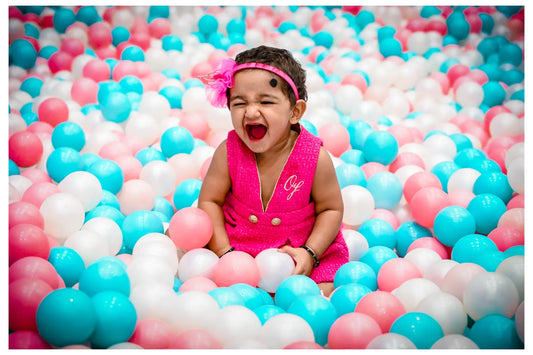 A baby girl in a pink dress laughs joyfully in a ball pit filled with pink, blue, and white balls.