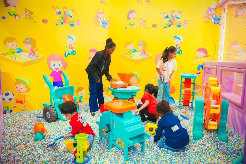 Children play in a colorful ball pit with toys, supervised by an adult.