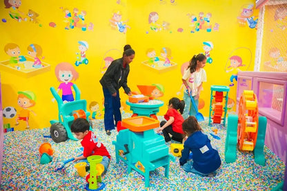 Children play in a colorful ball pit with toys, supervised by an adult.