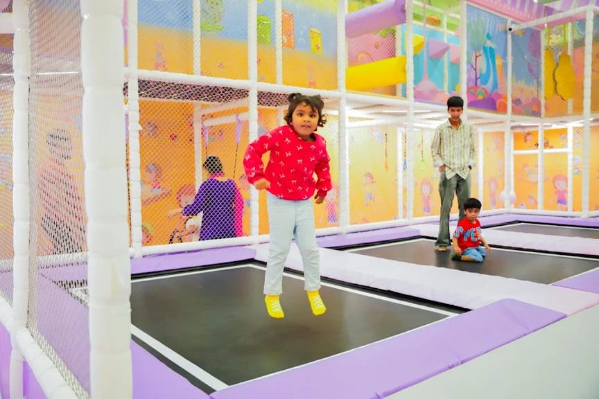 A young girl jumps on a trampoline in a colorful indoor play area, with other children and an adult in the background.