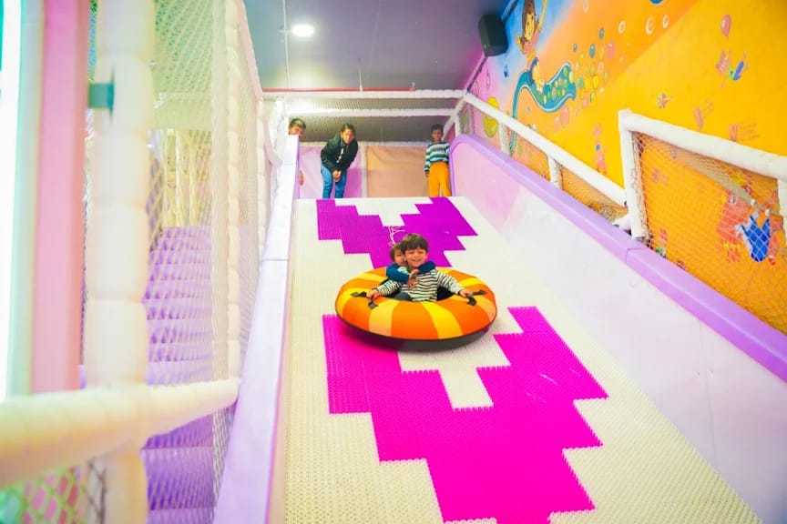 Two young children slide down a colorful tube slide in a play area.