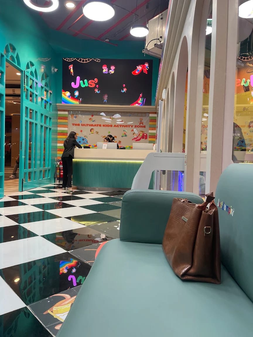 A reception area in a kids' activity zone with a checkered floor and a large screen displaying cartoon characters. A woman is at the counter, and a brown purse rests on a teal bench.