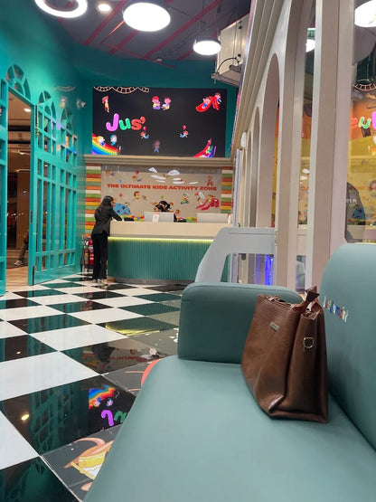 A reception area in a kids' activity zone with a checkered floor and a large screen displaying cartoon characters. A woman is at the counter, and a brown purse rests on a teal bench.
