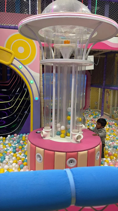 A young child plays in a ball pit next to a colorful, multi-level toy structure. The indoor play area is filled with soft balls of various colors.