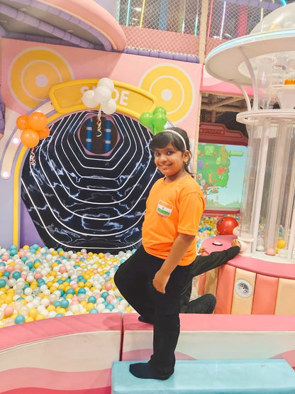A young girl in an orange shirt stands in a colorful ball pit area. She is smiling and looking at the camera.