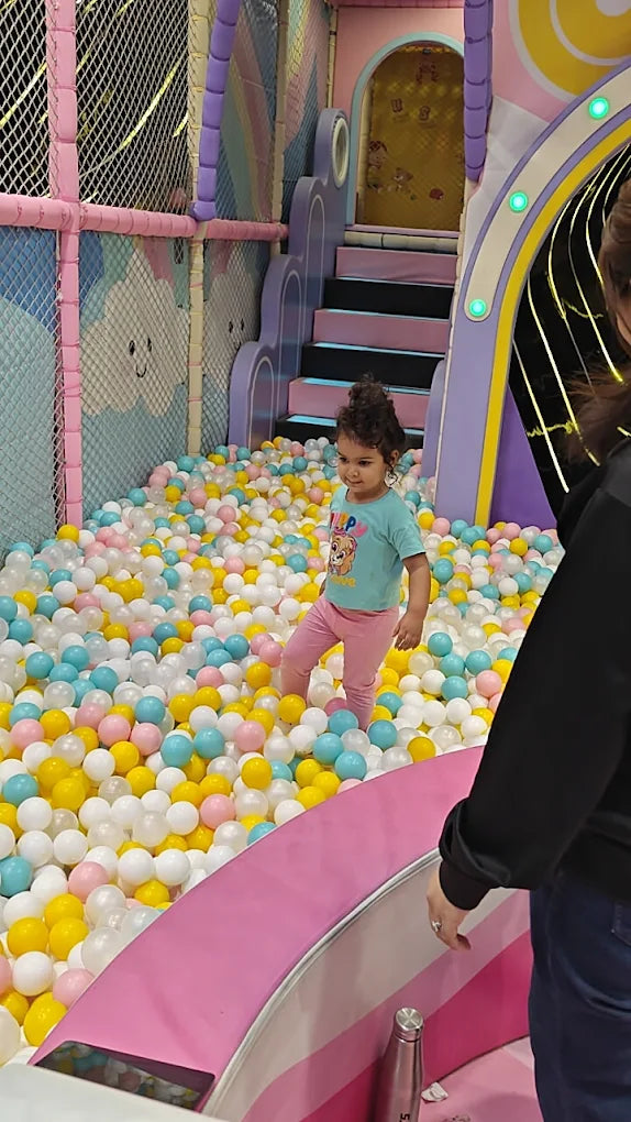 A young child plays in a ball pit at an indoor playground. The child is surrounded by colorful balls and a slide structure.