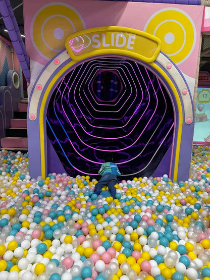 A child climbs into a colorful ball pit in front of a hexagonal tunnel slide with purple lights.