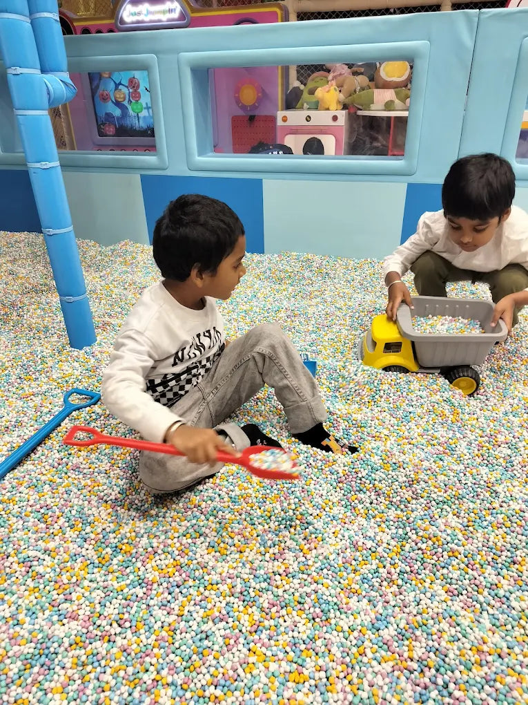 Two young boys play in a ball pit filled with colorful balls, one with a toy truck and the other with a red shovel.
