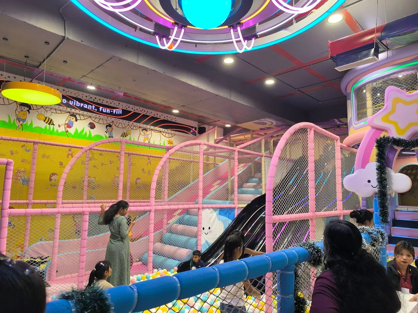 Children play in a colorful indoor playground with slides and ball pits, supervised by adults.