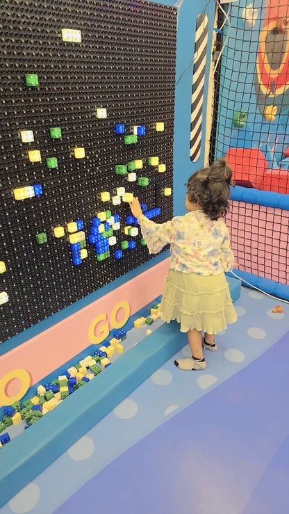 A young child plays with colorful blocks on a pegboard wall in a brightly colored indoor play area.