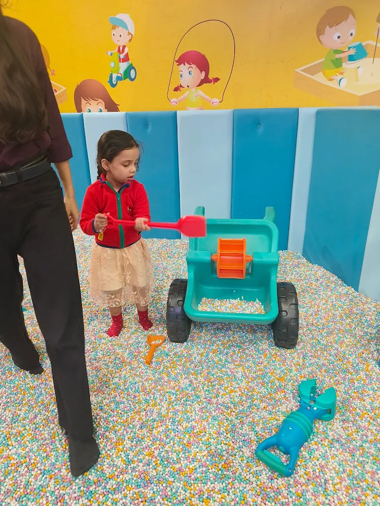 A young child plays with a toy wheelbarrow in a ball pit.