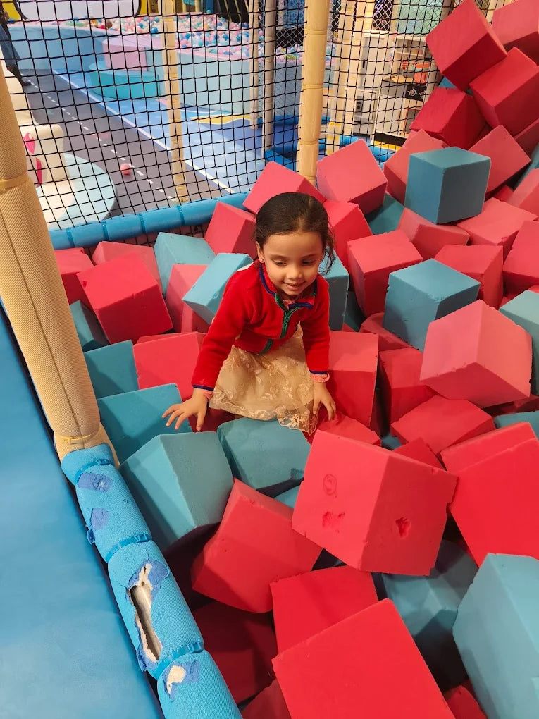 A young girl smiles while playing in a ball pit filled with red and blue foam blocks.