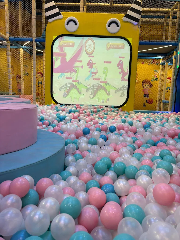 A ball pit filled with pink, blue, and white balls in front of a screen showing a dinosaur game.