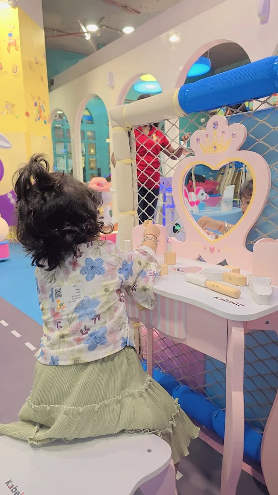 A young child plays at a pink vanity table in a colorful indoor play area.