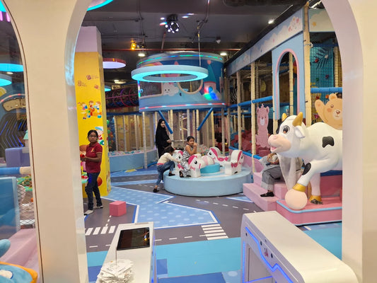 Children play in a colorful indoor playground with large animal figures. A young woman stands nearby.