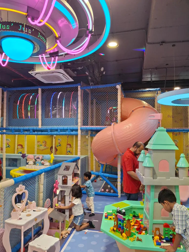 Children play in a colorful indoor playground with slides and toy houses, supervised by an adult.