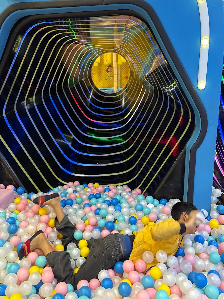 A young person lies in a ball pit, partially submerged, with a colorful tunnel above.