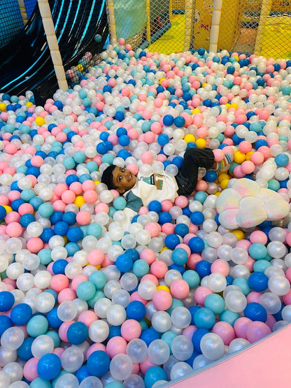 A young child lies in a ball pit filled with colorful balls, smiling at the camera.