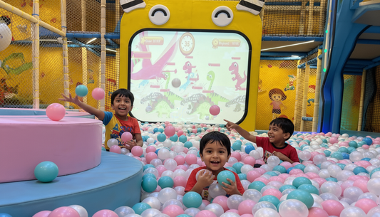 Three young boys play in a ball pit in front of a dinosaur-themed screen.