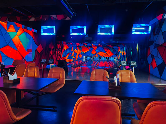 A vibrant bowling alley with neon lights, colorful geometric murals, and orange leather seating.
