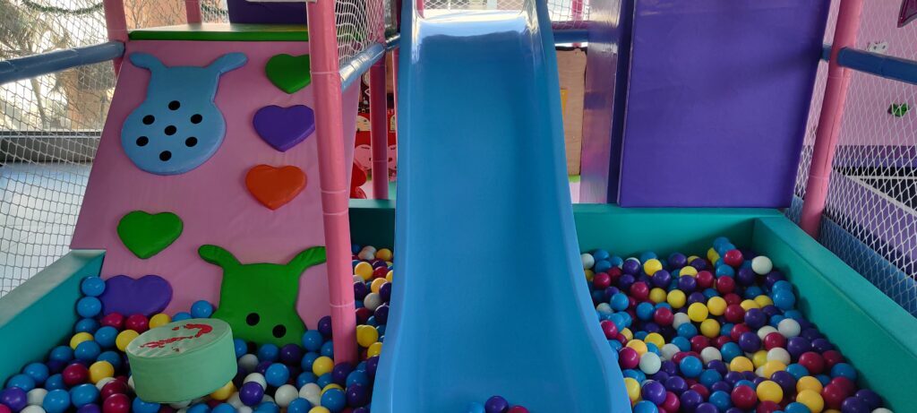 A colorful indoor playground with a blue slide and ball pit, featuring soft play shapes for young children.