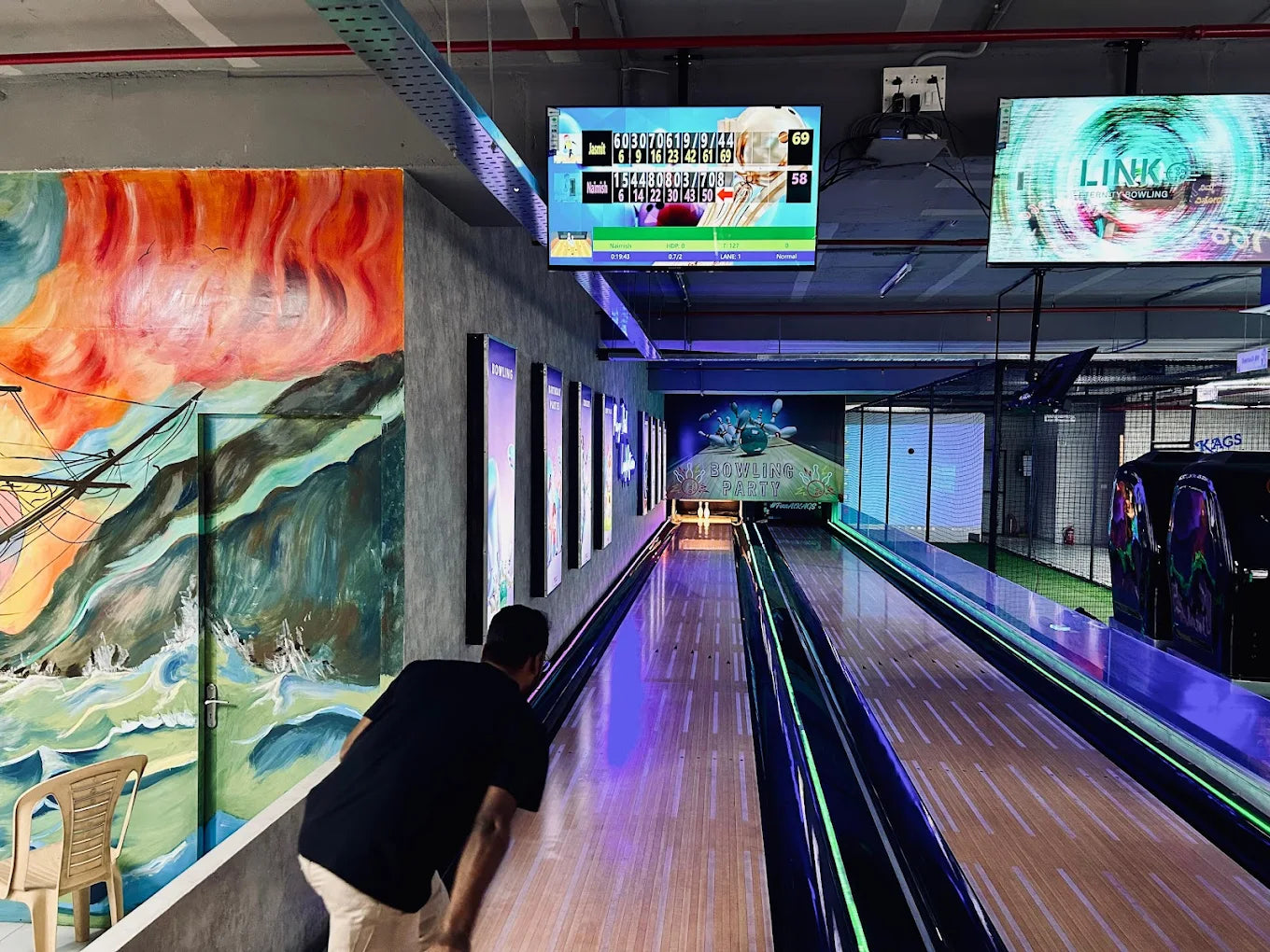 A man bowls in a brightly lit bowling alley with colorful murals and screens displaying scores.