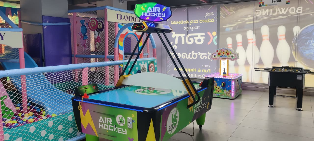 An air hockey table and foosball table in a brightly lit indoor arcade with a colorful soft play area for children.