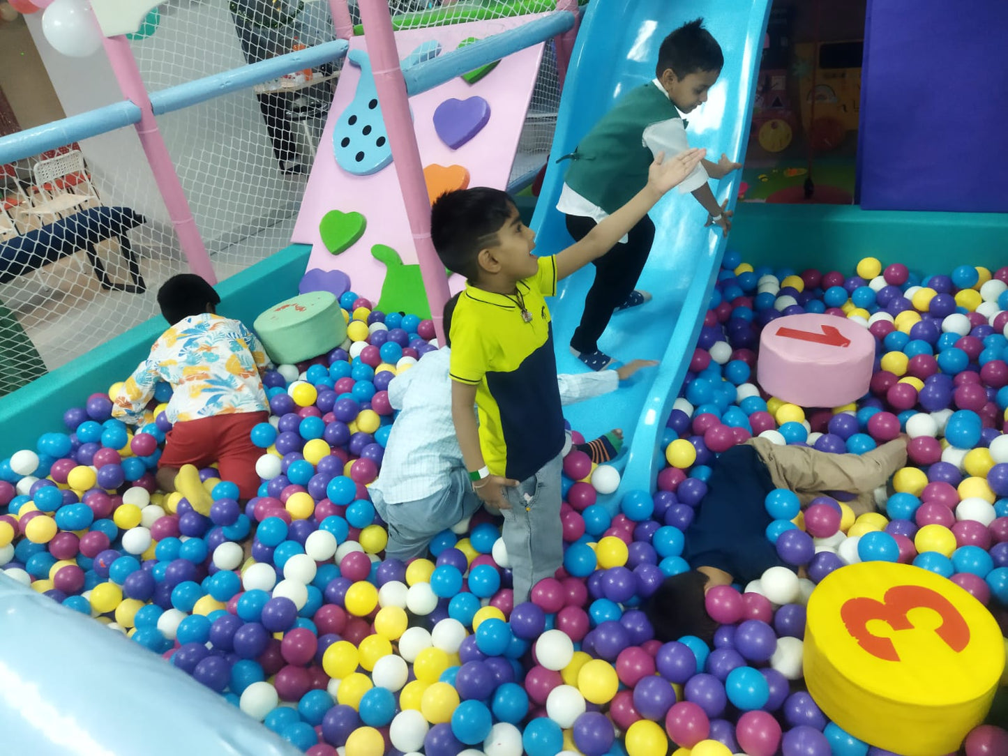 Children play in a ball pit with a blue slide. A boy in a green vest slides down as others play among the colorful balls.