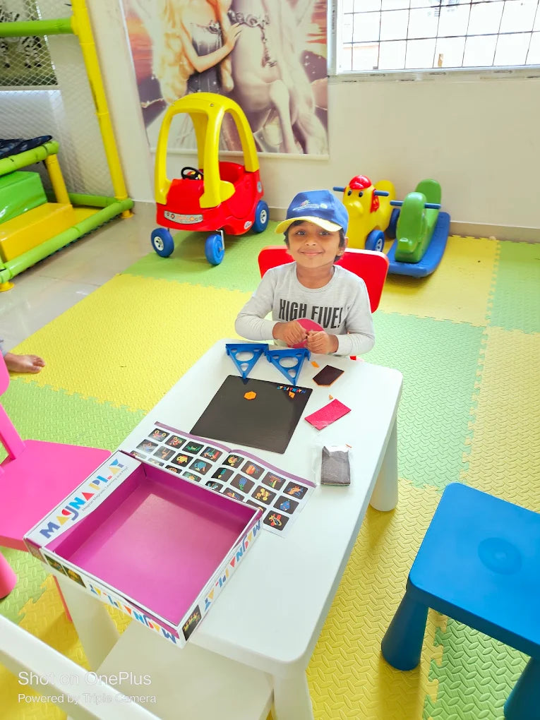 A young child sits at a table in a playroom, engaged in a magnetic play activity. Toys and colorful mats fill the background.