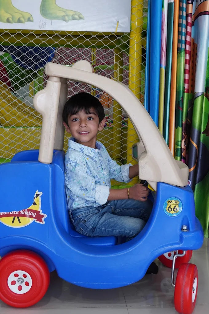 A young boy smiles while sitting in a blue toy car in a play area.