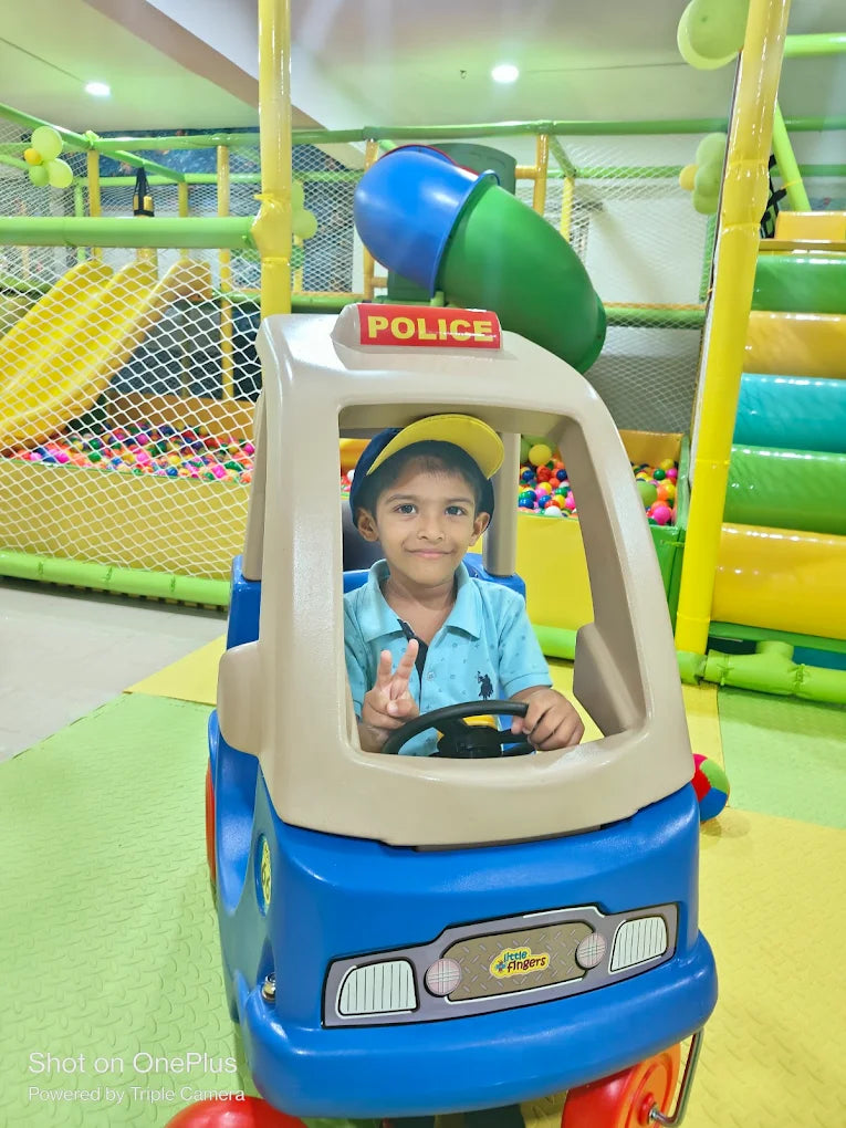 A young boy in a toy police car smiles while playing in an indoor playground.