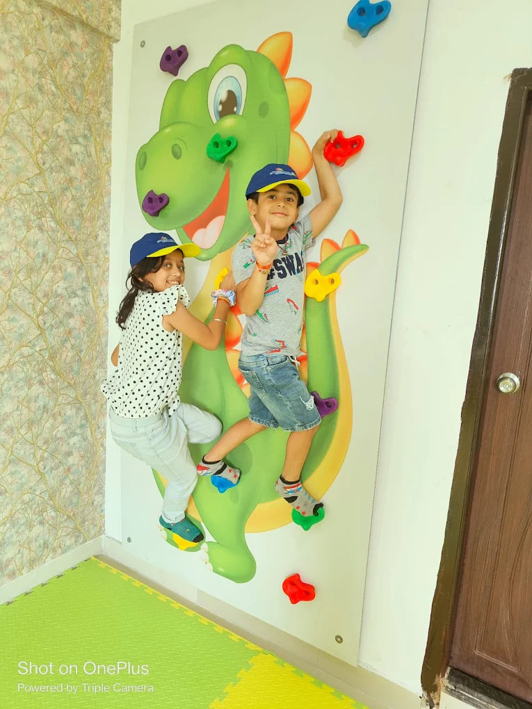 Two young children climb a colorful dragon-themed wall with colorful handholds.